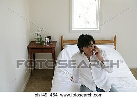 High angle view of a young man sitting on the bed and talking on a mobile phone View Large Photo Image Picture - High angle view of a young man sitting on the bed and talking on a mobile phone. Fotosearch