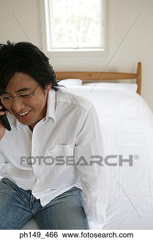 High angle view of a young man sitting on the bed and talking on a mobile phone View Large Photo Image Stock Photograph - High angle view of a young man sitting on the bed and talking on a mobile phone. Fotosearch