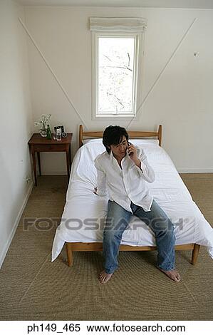 Stock Photography - High angle view of a young man sitting on the bed and talking on a mobile phone. Fotosearch