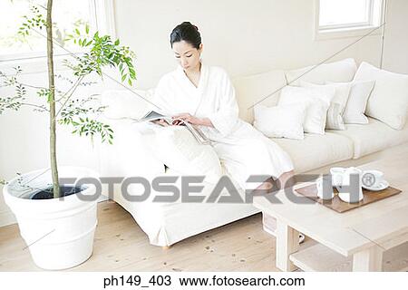 Stock Image - High angle view of a young woman sitting on a couch and reading a magazine. Fotosearch