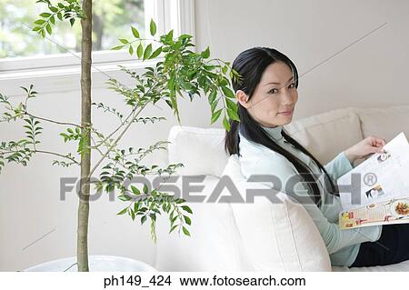 Portrait of a young woman sitting on a couch and holding a magazine View Large Photo Image Picture - Portrait of a young woman sitting on a couch and holding a magazine. Fotosearch