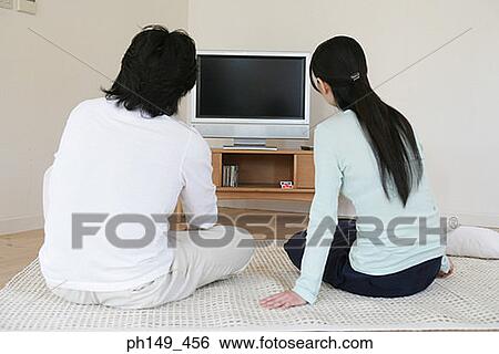 Rear view of a man and a woman sitting in front of a television View Large Photo Image Stock Photograph - Rear view of a man and a woman sitting in front of a television. Fotosearch