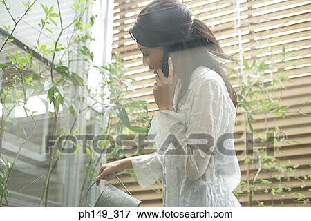 Side profile of a young woman talking on a mobile phone and watering plants View Large Photo Image Stock Photo - Side profile of a young woman talking on a mobile phone and watering plants. Fotosearch