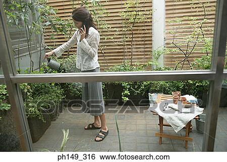 Side profile of a young woman talking on a mobile phone and watering plants View Large Photo Image Stock Photograph - Side profile of a young woman talking on a mobile phone and watering plants. Fotosearch