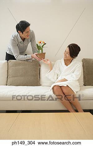 Picture - Young man giving a potted plant to a young woman sitting on a couch. Fotosearch