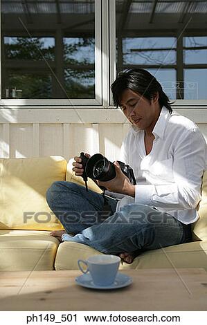 Young man sitting on a couch and holding a camera View Large Photo Image Stock Image - Young man sitting on a couch and holding a camera. Fotosearch