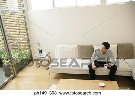 Young man sitting on a couch and holding a cup of coffee View Large Photo Image Stock Photo - Young man sitting on a couch and holding a cup of coffee. Fotosearch
