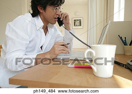 Picture - Young man using a laptop and talking on the telephone. Fotosearch