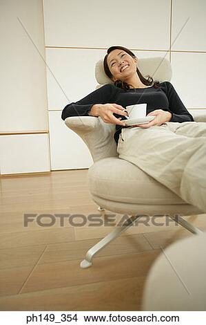 Young woman holding a cup of coffee and a saucer in an armchair View Large Photo Image Picture - Young woman holding a cup of coffee and a saucer in an armchair. Fotosearch