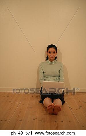 Stock Image - Young woman leaning against a wall with a laptop on her lap. Fotosearch