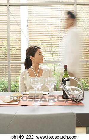 Young woman sitting at the dining table with a man walking in the background View Large Photo Image Stock Image - Young woman sitting at the dining table with a man walking in the background. Fotosearch