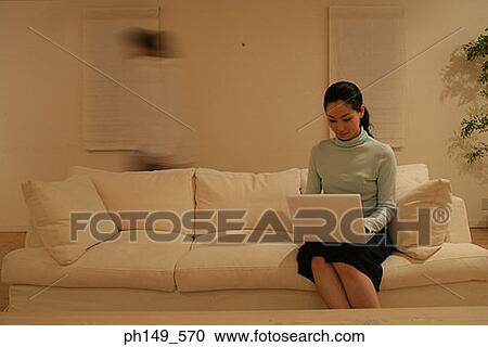 Young woman sitting on a couch and using a laptop View Large Photo Image Stock Image - Young woman sitting on a couch and using a laptop. Fotosearch