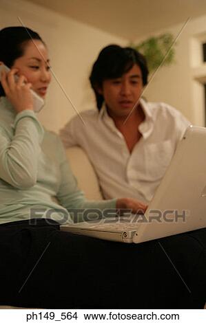 Young woman talking on a mobile phone and using a laptop with a young man sitting beside her View Large Photo Image Picture - Young woman talking on a mobile phone and using a laptop with a young man sitting beside her. Fotosearch