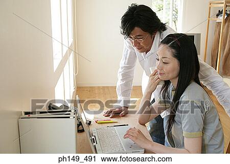 Stock Image - Young woman using a laptop with a young man standing beside her. Fotosearch