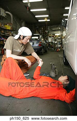 Stock Photography - Close-up of a female auto mechanic repairing a car with another mechanic crouching beside her. Fotosearch