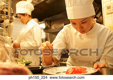 Picture - Close-up of a female chef dripping sauce on a plate with her colleague standing behind her. Fotosearch