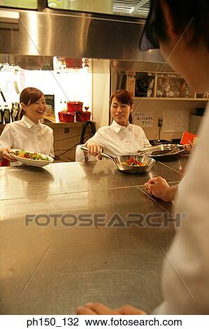 Two waitresses standing at a kitchen counter and serving food View Large Photo Image Stock Image - Two waitresses standing at a kitchen counter and serving food. Fotosearch