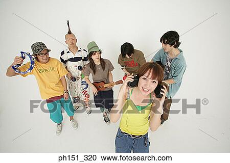 Stock Image - Portrait of a young woman holding headphones with her friends playing musical instruments behind her. Fotosearch