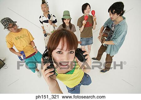 Portrait of a young woman holding headphones with her friends playing musical instruments behind her View Large Photo Image Stock Image - Portrait of a young woman holding headphones with her friends playing musical instruments behind her. Fotosearch