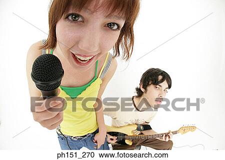 Portrait of a young woman singing into a microphone with a young man playing the guitar behind her View Large Photo Image Stock Image - Portrait of a young woman singing into a microphone with a young man playing the guitar behind her. Fotosearch