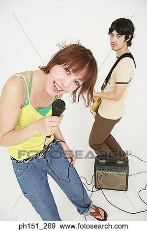 Portrait of a young woman singing into a microphone with a young man playing the guitar behind her View Large Photo Image Stock Photo - Portrait of a young woman singing into a microphone with a young man playing the guitar behind her. Fotosearch