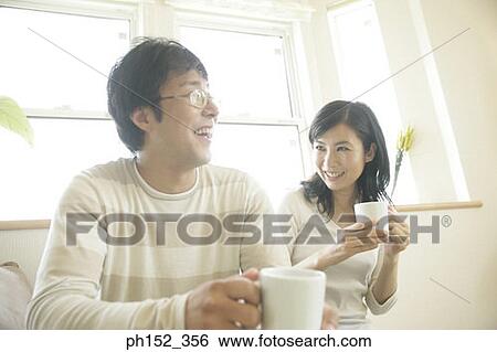Close-up of a mid adult couple sitting on a couch and holding cups of coffee View Large Photo Image Stock Photograph - Close-up of a mid adult couple sitting on a couch and holding cups of coffee. Fotosearch