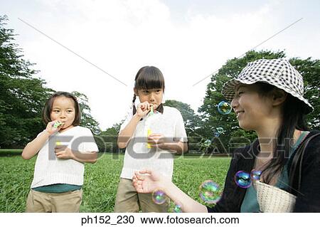 Stock Image - Close-up of a mid adult woman and her two daughters blowing bubbles in the park. Fotosearch