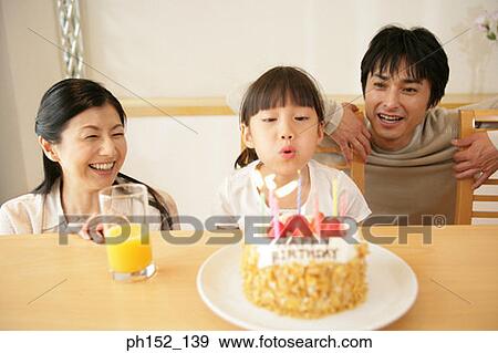 Stock Photo - Girl blowing out candles on a birthday cake with their parents beside her. Fotosearch