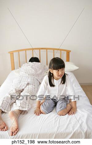 Stock Photo - Girl sitting on the bed with her father sleeping beside her. Fotosearch