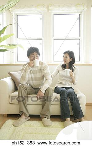 Stock Image - Mid adult couple sitting on a couch and holding cups of coffee. Fotosearch