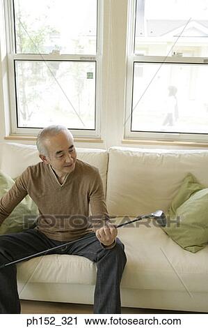 Stock Image - Senior man sitting on a couch and holding a golf club. Fotosearch
