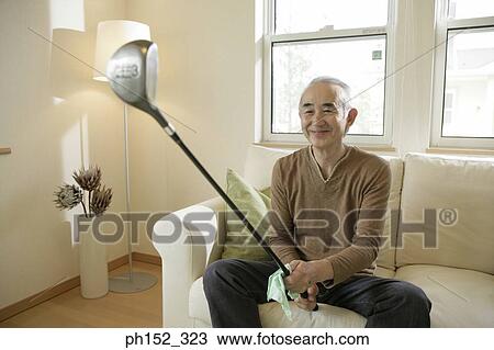 Stock Image - Senior man sitting on a couch and holding a golf club. Fotosearch