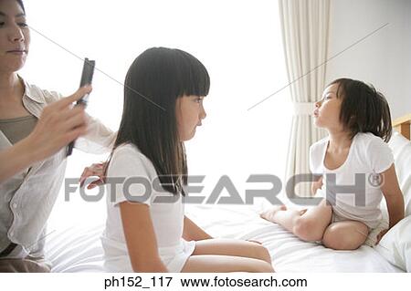 Stock Photo - Side profile of a girl and her sister sitting on the bed with their mother brushing her hair. Fotosearch