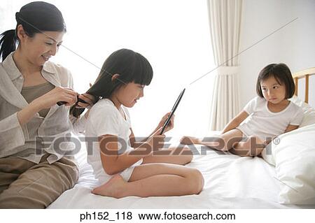 Stock Photo - Side profile of a girl and her sister sitting on the bed with their mother brushing her hair. Fotosearch