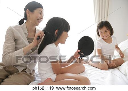 Stock Photo - Side profile of a girl and her sister sitting on the bed with their mother brushing her hair. Fotosearch