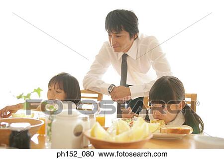 Two girls sitting at the dining table with their father leaning against their chairs behind them View Large Photo Image Stock Image - Two girls sitting at the dining table with their father leaning against their chairs behind them. Fotosearch