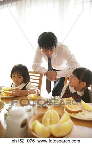 Stock Photograph - Two girls sitting at the dining table with their father leaning against their chairs. Fotosearch