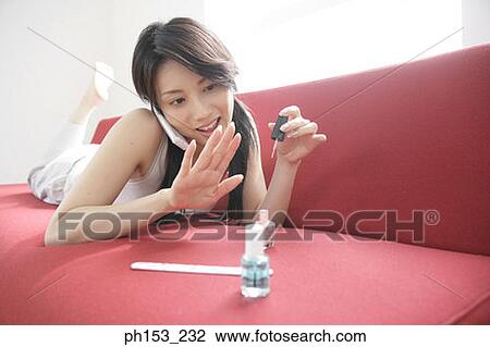 Stock Image - Close-up of a young woman applying nail polish on her fingernails and talking on a mobile phone. Fotosearch