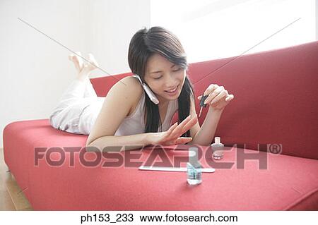 Stock Image - Close-up of a young woman applying nail polish on her fingernails and talking on a mobile phone. Fotosearch