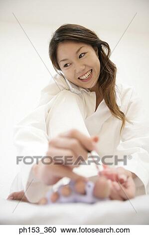 Stock Image - Close-up of a young woman applying nail polish on her toenails and talking on a mobile phone. Fotosearch