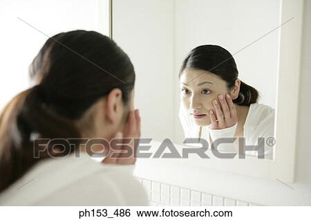 Close-up of a young woman looking into a mirror and touching her face View Large Photo Image Stock Photograph - Close-up of a young woman looking into a mirror and touching her face. Fotosearch