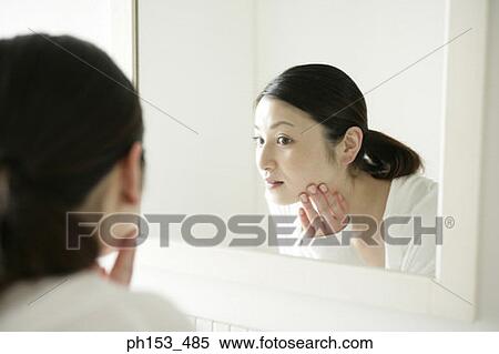 Close-up of a young woman looking into a mirror and touching her face View Large Photo Image Stock Photography - Close-up of a young woman looking into a mirror and touching her face. Fotosearch