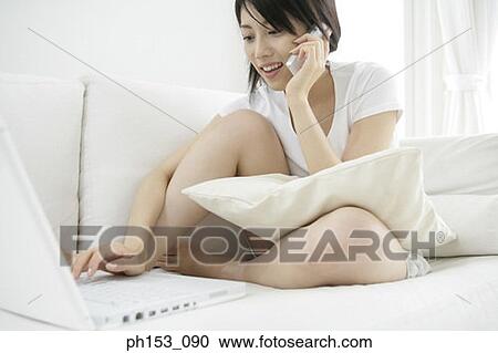 Close-up of a young woman sitting on a couch and using a laptop while talking on a mobile phone View Large Photo Image Stock Image - Close-up of a young woman sitting on a couch and using a laptop while talking on a mobile phone. Fotosearch