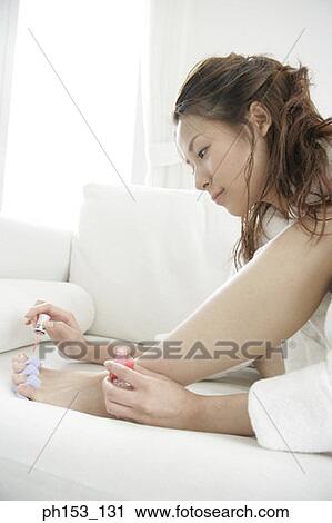 Stock Image - Close-up of a young woman sitting on a couch and applying nail polish. Fotosearch