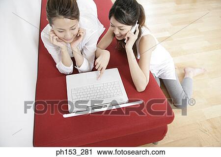 High angle view of a young woman lying on a couch with another young woman using a laptop and talking on a mobile phone beside her View Large Photo Image Stock Image - High angle view of a young woman lying on a couch with another young woman using a laptop and talking on a mobile phone beside her. Fotosearch