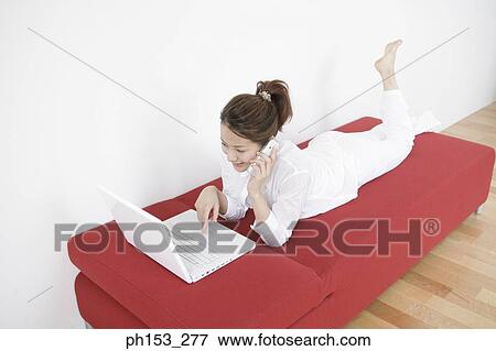 High angle view of a young woman lying on a couch and using a laptop while talking on a mobile phone View Large Photo Image Stock Photo - High angle view of a young woman lying on a couch and using a laptop while talking on a mobile phone. Fotosearch
