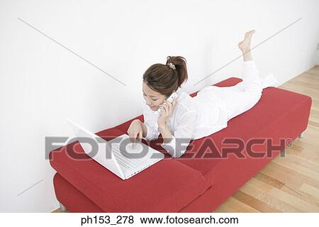 High angle view of a young woman lying on a couch and using a laptop while talking on a mobile phone View Large Photo Image Stock Photo - High angle view of a young woman lying on a couch and using a laptop while talking on a mobile phone. Fotosearch