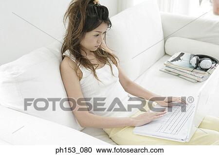 Stock Image - High angle view of a young woman reclining on a couch and using a laptop. Fotosearch