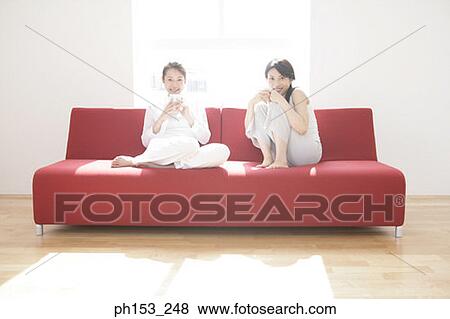 Portrait of two young women sitting on a couch and holding cups of coffee View Large Photo Image Stock Photo - Portrait of two young women sitting on a couch and holding cups of coffee. Fotosearch