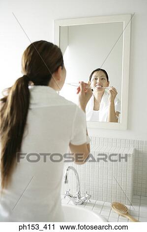 Stock Image - Rear view of a young woman brushing her teeth in front of a mirror. Fotosearch